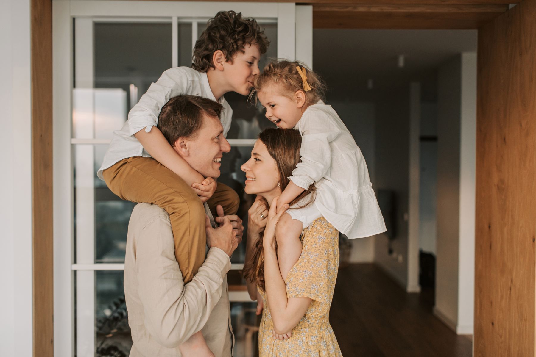 Two parents at home laughing with their young son and daughter on their shoulders